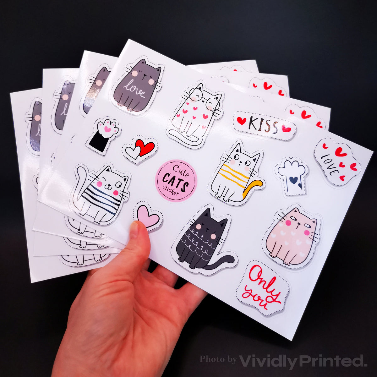 A hand holding sticker sheets with cartoon cats and hearts